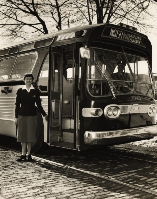 A woman in a TTC uniform consisting of a dark jacket and knee-length skirt stands beside the open door of a bus.