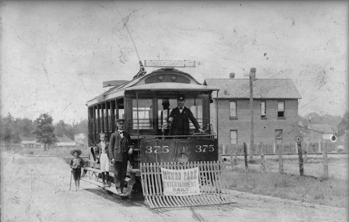 A small electric streetcar. ON the front is a banner reading "Munro Park, entertainment daily." A man in a driver's uniform stands on the the step, and another in the driver's platform at the front.