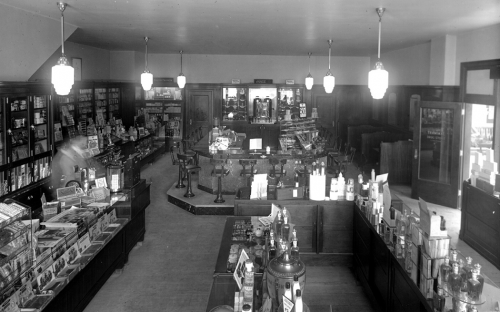 Inside a store containing many wooden shelves full of goods. In the background is a lunch counter surrounded by high chairs. 