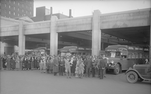 Several buses are parked in a half-open garage. Numerous people are posed in front of them and are looking at the camera.