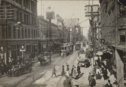 A busy street lined with office buildings and stores. There is a lot of vehicle and pedestrian traffic.