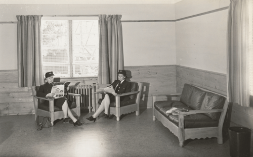 Two uniformed women seated by window reading magazines.