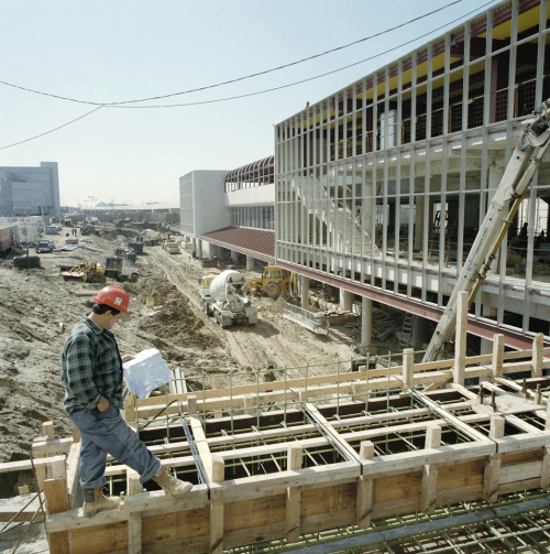 Man walking along elevated structure towards an unfinished 3-strorey building. Unfinished driveway and bus waiting area in background.