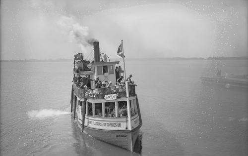 A small wooden ferry with an enclosed lower deck and people standing on the open upper deck.