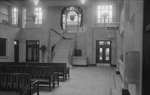 A waiting room filled with long wooden benches. In the background, a staircase leads up to a landing with a large arched window with the Grey Coach crest in it. 