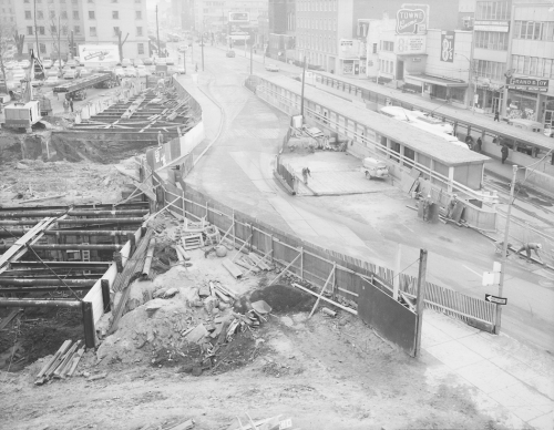 Excavation for tunnel and bus platforms in street. Storefronts line the far side of the street.