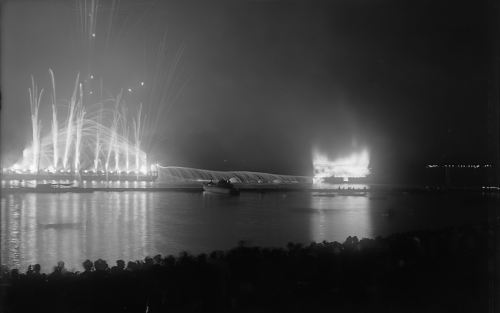 At night, a ship engulfed in flames floats on the water. There are fireworks going off beside it.