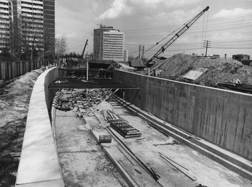Construction of subway trackbed structure. Background shows crane beside tunnel structure, apartment building, and office tower.