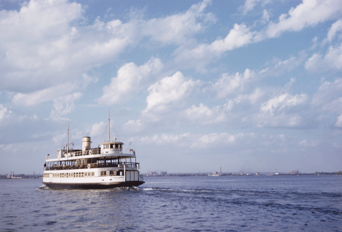 A large wooden ferry painted white chugs across blue water under a sky filled with fluffy white clouds.