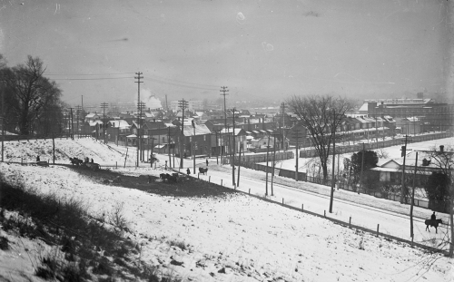 Looking down a hill to a road and houses beyond, all covered in snow.