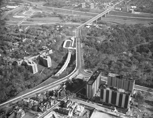 Apartment buildings and other buildings in foreground. Prince Edward Viaduct in background. Middle shows cars on Bloor Street and station structure