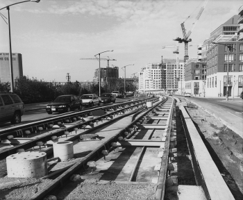 Newly installed track in unfinished right or way with motor vehicle traffic on roadway. Background shows condominiums.