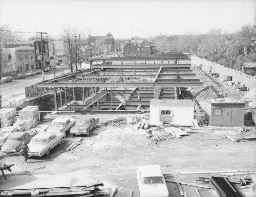 Structural beams erected prior to station construction. Foreground shows vehicles and workers trailers, background shows storefront lining street.