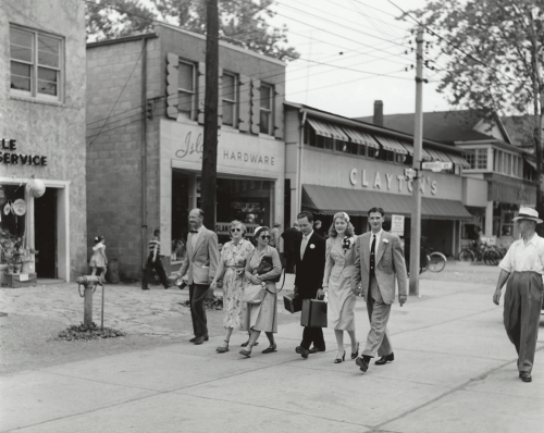 People are walking down a street with stores in the background. The store signs read "Island Hardware" and "Clayton's."