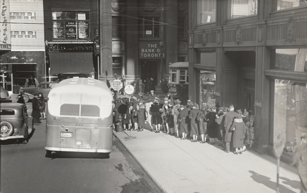 Men and women lined up to get on bus. Background show storefronts.