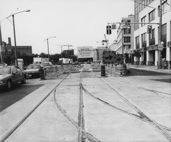 Looking along installed track on right of way with road traffic. Sign advertising Queens Quay LRT.