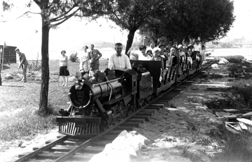 People are riding a miniature train with a small locomotive and open two-person cars.