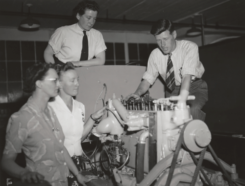 Three women standing about bus motor with instructor lecturing.