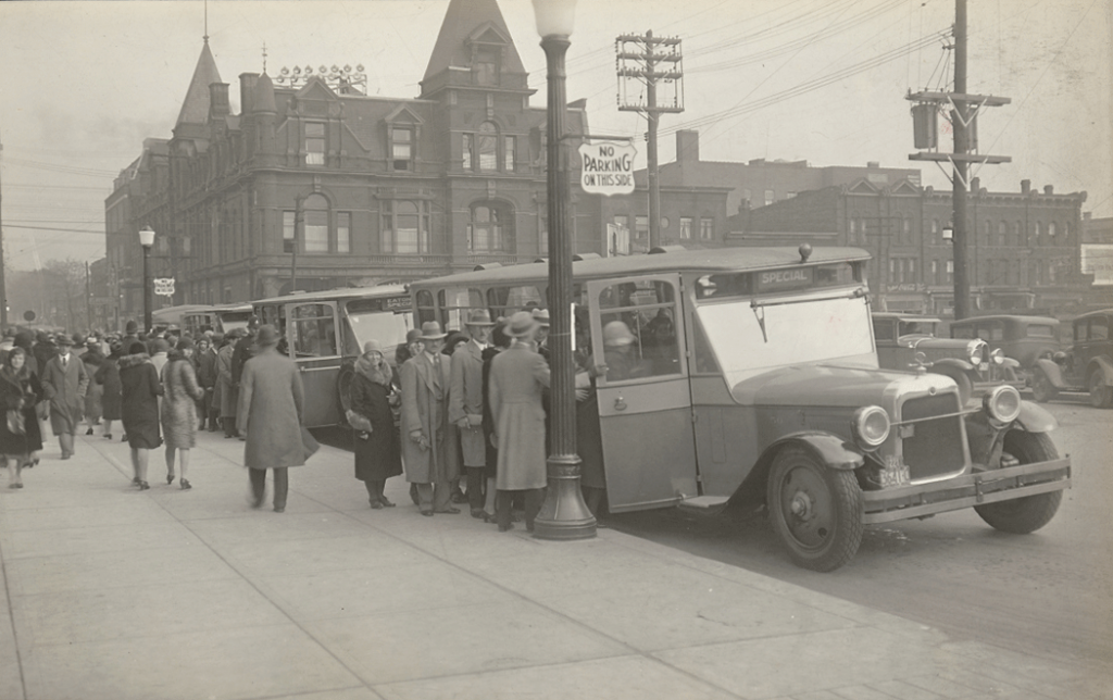 Men and women lined up to board bus. Background contains brick buildings.