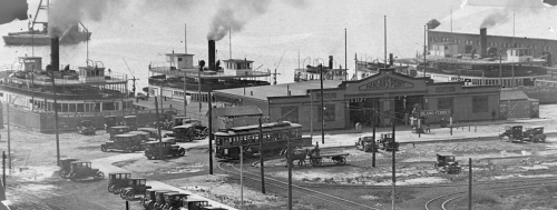 A low building on the waterfront, with two large open doors and a peaked roof with a sign saying "Hanlan's Point." Behind it in the water are two ferries with smoking smokestacks.