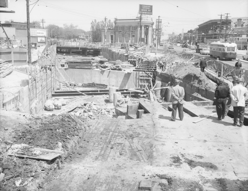 Construction workers looking toward excavated area construction and traffic along Bloor Street. Background shows bank and storefronts along road.