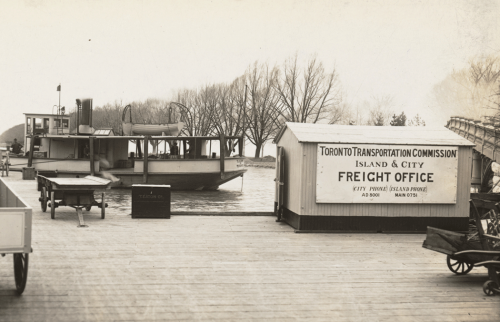 A wide wooden dock with a ferry at the side and several carts waiting for freight. There is a small building with a sign that reads "Island and City freight office."