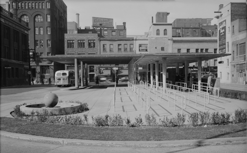 Foreground shows passenger waiting area and bus boarding location. Background shows station and buildings.