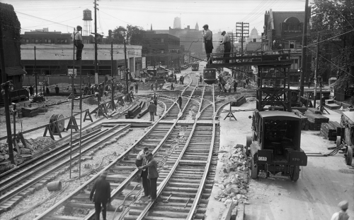 Workers are installing two parallel sets of streetcar tracks on the road. Other workers are standing in a crane lift and on a tall ladder to install overhead wires.