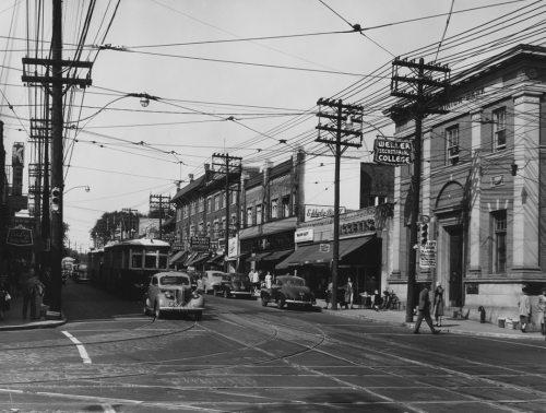 A busy street with cars and a streetcar. Lining the street are stores and services, including Weller Secretarial College.