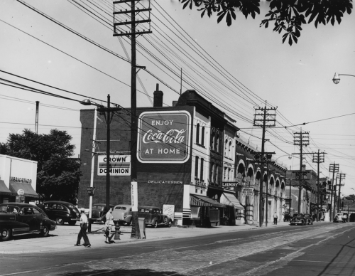 A parking lot with a row of stores behind it, and a billboard that reads, "Enjoy Coca-Cola at home."