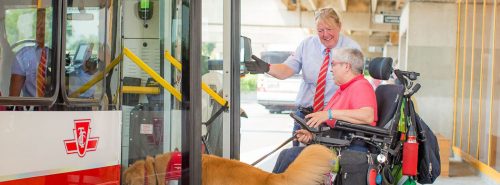 A man sits in a wheelchair and holds the leash of a golden retriever dog. A smiling TTC driver gestures at him to enter to open bus doors.