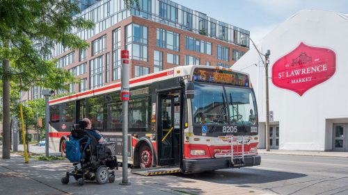 A man in a wheelchair sits in front of a ramp leading onto a TTC bus. Behind the bus is a sign for the St. Lawrence Market.