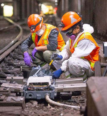 People in hardhats and protective masks weld electronic components together in a subway tunnel.