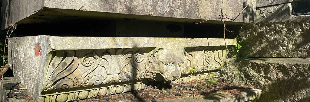 A large rectangular relic stone with ornate patterns and shapes with a small lions head. The stone is partially covered in light green moss and shows signs of weathering and corrosion.