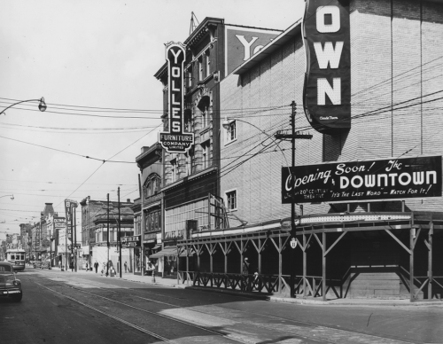 The exterior and sign of the Yolles Furniture Company and a fenced-off lot with a sign saying "Coming Soon, The Downtown."