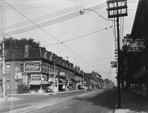 A row of identical three-storey brick stores. On the corner is a store with awnings over the entrance and a large Coca-Cola sign on the side.