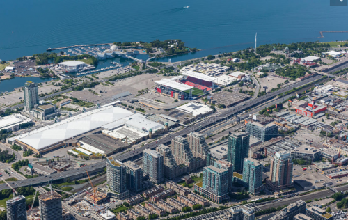 Exhibition Place grounds seen from above, looking south west toward Lake Ontario. The image shows large, flat exhibition buildings, parking lots and roads, surrounded by the highway and rail corridor to the north