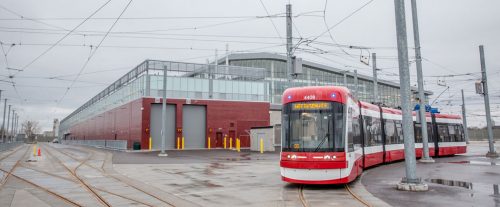 A long red streetcar stands in front of a modern brick and glass building in the rain.