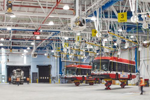 Two TTC buses are held off the ground by jacks and ropes in a large white garage.