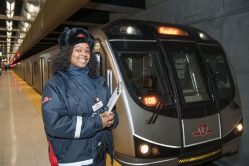 A smiling Black woman in a TTC coat and winter hat stands beside a subway train.