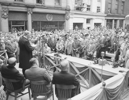 A man stands at a microphone on a platform set up on the street. Other people are sitting behind him, and there is a crowd in front of the platform.