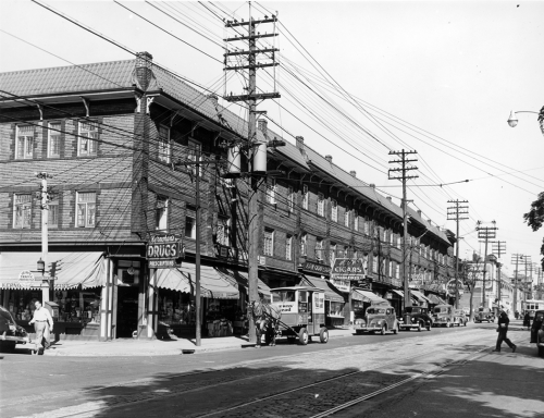 A long three-storey building containing many stores. A Brown's Bread delivery wagon pulled by a horse is parked at the curb.