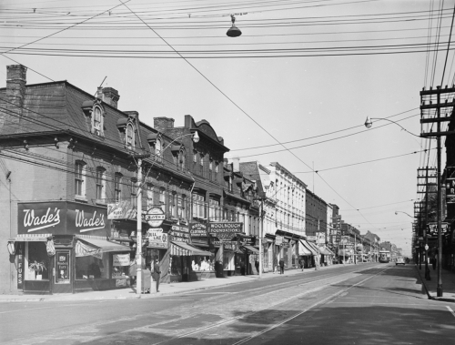 Many stores on one side of a street, include Wade's Furs and Midtown Clothing.