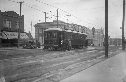 People line up to bard a streetcar in the middle of the street. In the background are stores, including a W. Davies butcher shop.