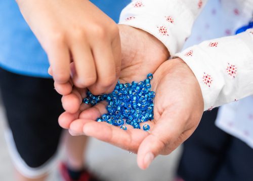 Cupped hands holding small blue beads; another person's hand reaching in to pick up a few beads