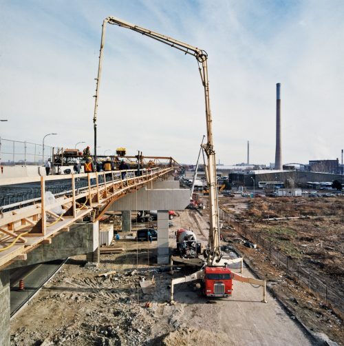 Red concrete truck has long crane-like arm that lifts hose up to deck of elevated expressway.