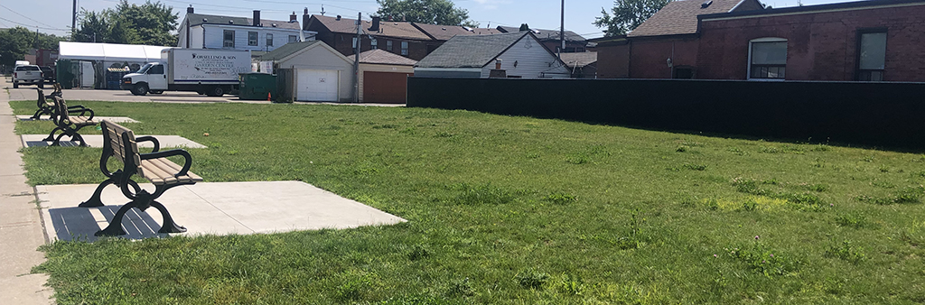 A photograph of the location for the new park at 261 Nairn Avenue on a sunny day. The image shows an open area with grass and three park benches on a concrete pad with residential homes surrounding the area.
