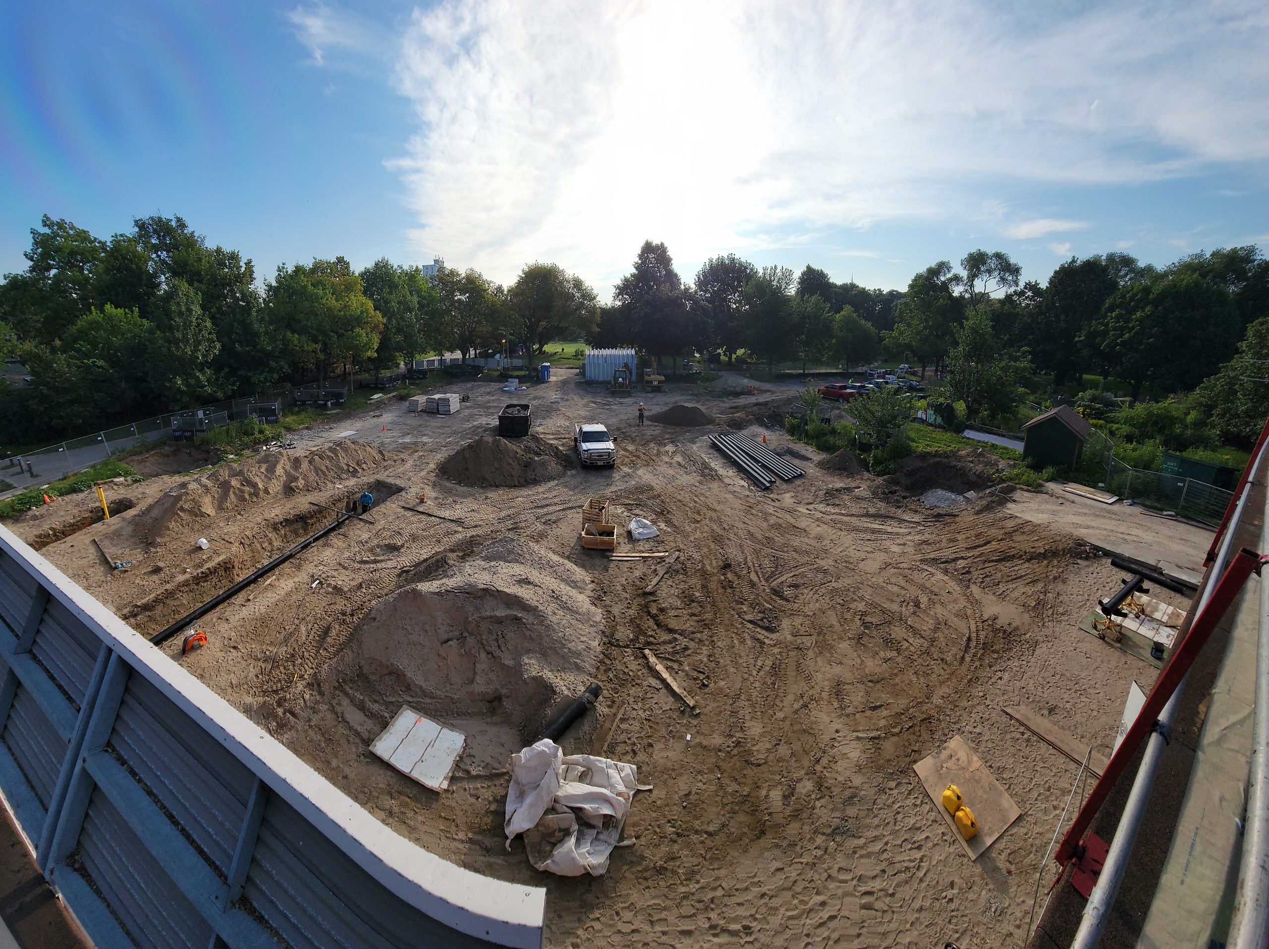 Photo shows the exterior construction site, from the roof of the Dufferin Grove Clubhouse, looking east over the old ice rink area and green space. The constructions site is mostly dirt and piles of gravel, with one worker in a dug-out trench, working with black piping. Wood, piping, and other construction debris are scattered around the site. A white pickup truck is parked in the middle of the site.