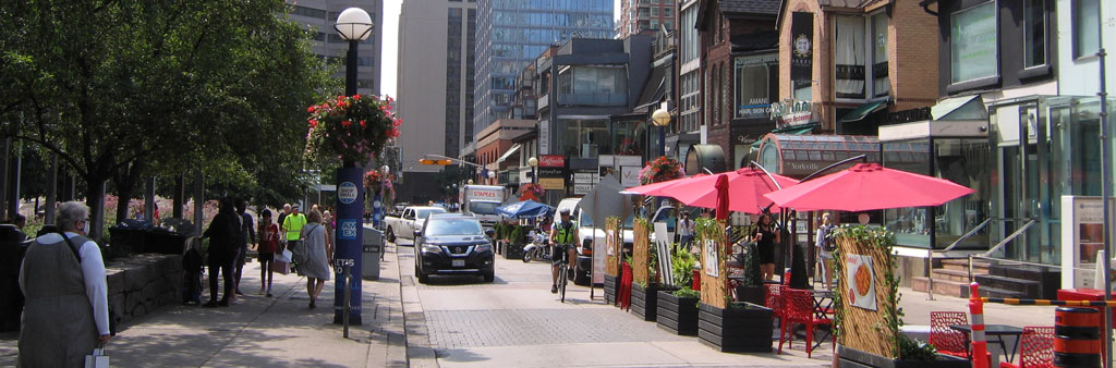 Cumberland Street in the Village of Yorkville, showing dining patios along the road and pedestrians walking next to a park