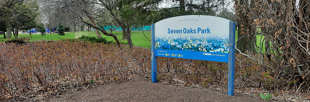 An image of the park sign at the entrance of Seven Oaks Park. Mature trees and plants surround the sign.